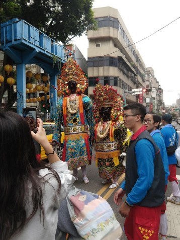 Two people in elaborate costumes with masks and fancy headdresses. One is tall and thin, one is short and squat.