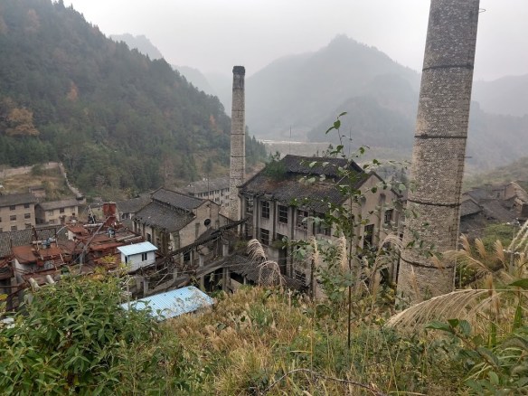 Two smokestacks and some very abandoned-looking buildings with mist-shrouded hills behind them.