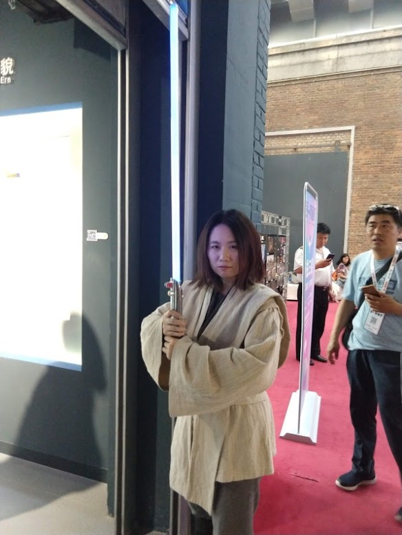 A Chinese woman in Jedi robes, holding a lightsaber upright.