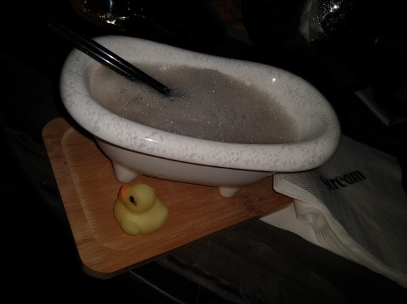 A miniature bathtub filled with foam. Two straws are sticking out. The bathtub rests on a wooden cutting board, which also holds a rubber duckie.
