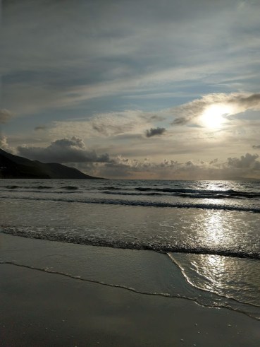 The sun low in the sky, partially obscured by clouds, over a beach.