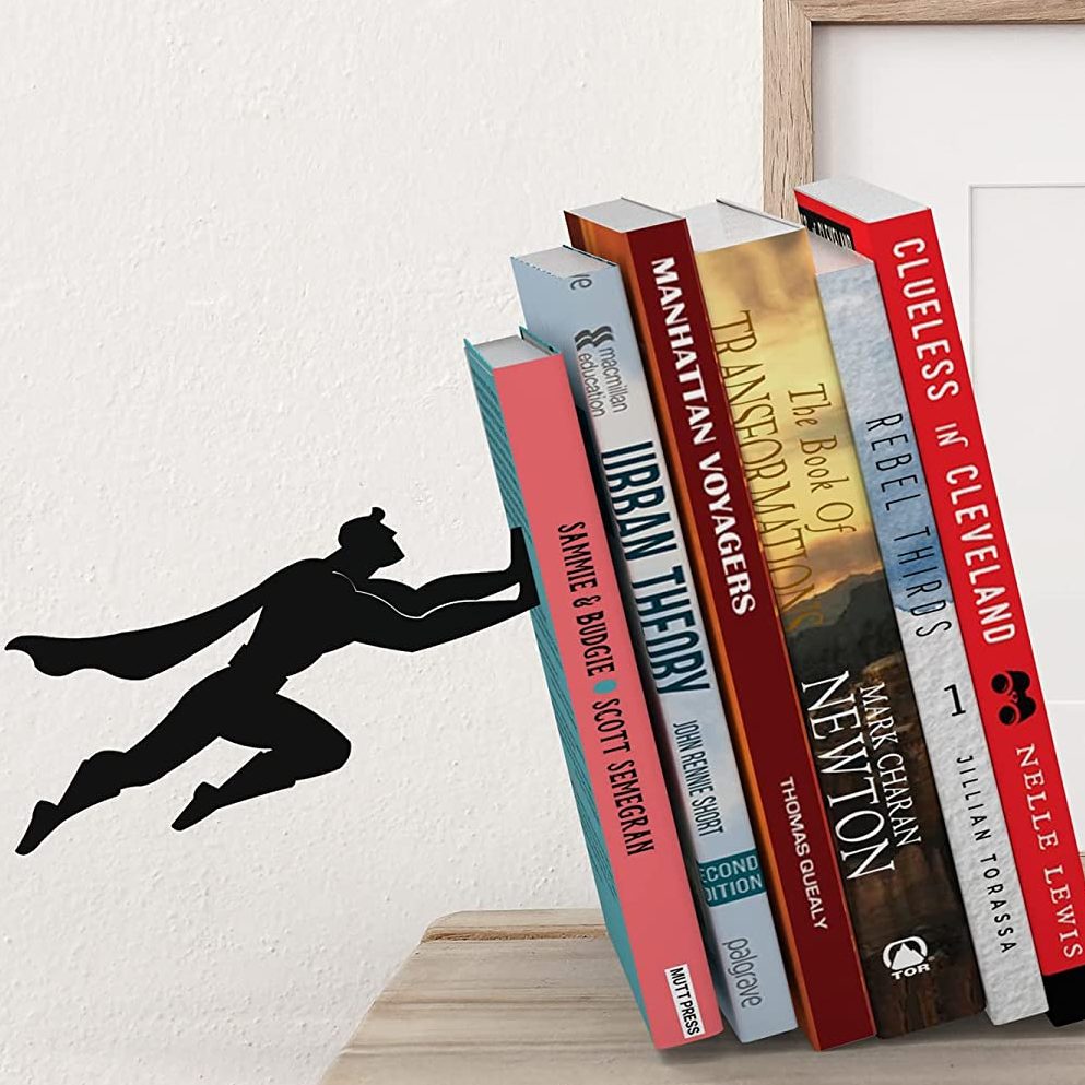 A silhouette of Superman appears to be flying and pushing against a stack of books to keep them from falling over. The books are leaning slightly to the side. 