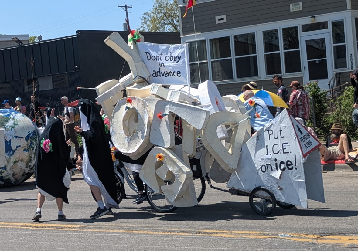A couple of people in penguin costumes are next to a bike loaded down with papier-mache letters and signs saying "Don't Obey in Advance!" and "Put ICE on ice!"